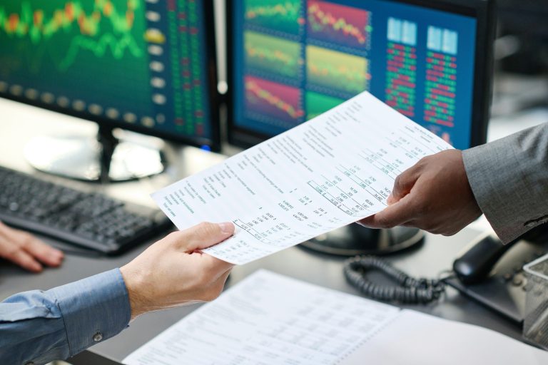 Börsenbriefe Zwei Personen übergeben sich Dokumente vor Monitoren mit Finanzgrafiken.